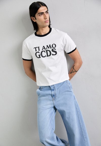 Bela skrajšana majica z črnim ovratnikom, na kateri je v debelih črnih črkah napis "TI AMO GCDS". Svetlo modri široki kavbojki.