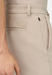 Beige byxor med en slät textur, med en BOGGI-etikett, en dragkedjeficka på sidan och en platt framfickdetail.