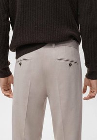 Pantaloni grigi chiari su misura con pieghe, due tasche posteriori, accentuati da bottoni scuri. Indossati con un maglione a coste scuro.