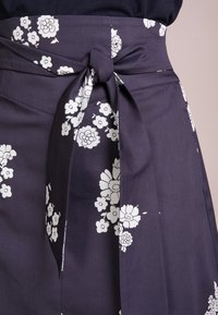 Tummansininen kangashame, jossa on valkoisia kukkakuoseja, sidottu vyötärölle yhteensopivalla kankaisella vyöllä. Sujuva rakenne ja ryhdikäs muotoilu.