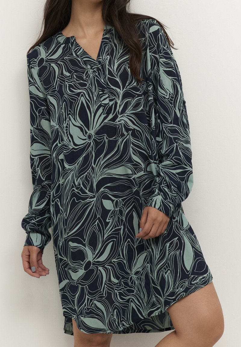 Femme portant une robe bleu marine à manches longues avec de grands motifs floraux vert clair et un col en V.