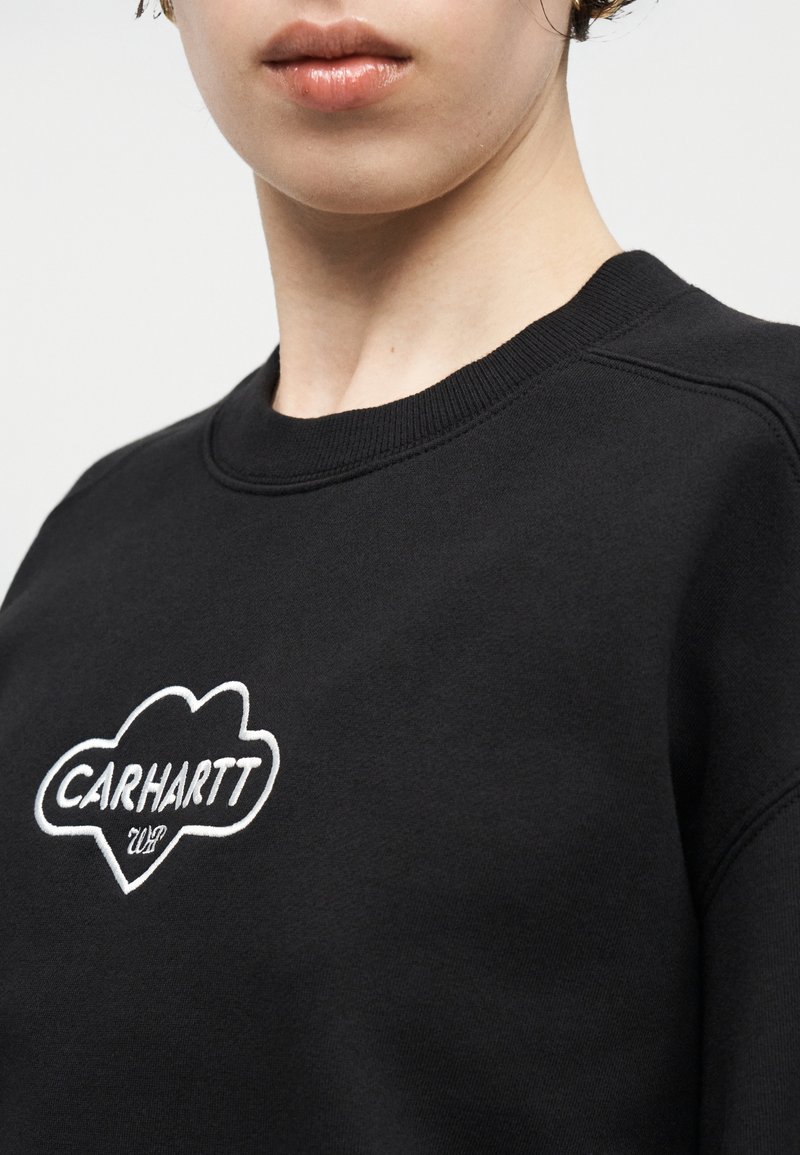 Primo piano di una persona che indossa una felpa nera con un logo ricamato bianco "CARHARTT WIP" sul petto.