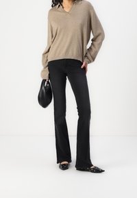 Pull en tricot beige avec un col en V, jean noir évasé et chaussures plates noires avec des œillets, tenant un sac noir.