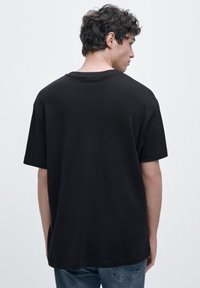 Svart bomullst-shirt med korta ärmar, avslappnad passform och rund halsringning. Mjuk textur, utan prydnader, visas bakifrån.