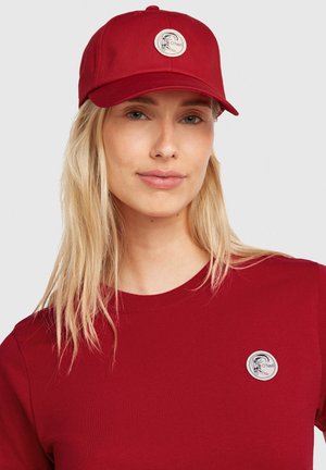 Punainen lippis, jossa on kaareva lippa ja pyöreä merkki, jossa on logo. Malli käyttää vastaavan punaista pyöreäkaulaista paitaa.