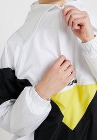 Biała i czarna wiatrówka z żółtym akcentem, z wysokim kołnierzem, zapięciem na półzamek z przodu i elastycznymi mankietami. Na piersi widoczne logo.