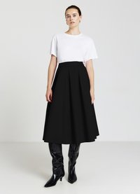 Gonna nera a lunghezza ginocchio con pieghe e vita alta, abbinata a una t-shirt bianca attillata. Modella che indossa stivali neri lucidi fino al ginocchio.