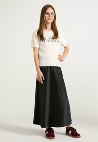 Cremefarbener, fusseliger Kurzarm-Pullover mit "Voilà" in Grau, kombiniert mit einem langen schwarzen Satrok und bordeauxfarbenen Sneakers mit strukturiertem Finish.