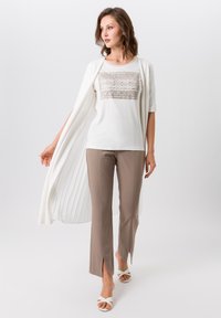 Witte top met metalen ornament, licht crèmekleurige lange cardigan, beige cropped broek met zijsplitten en witte sandalen met bandjes.