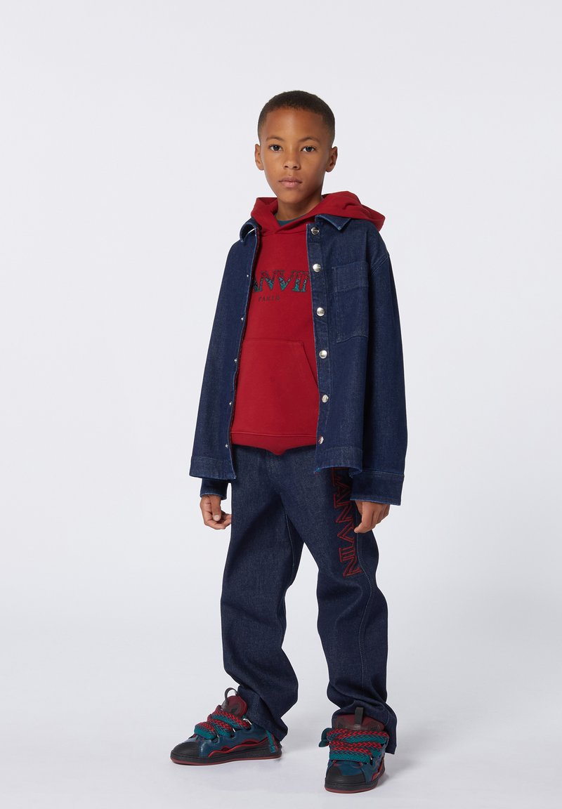 Giubbotto di jeans, felpa rossa con logo, pantaloni in denim scuro, sneakers multicolori con accenti testurizzati. Outfit casual con design a strati.