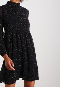 Black dress with a high ruffled neck, long sheer sleeves, and a flowing skirt, featuring white polka dots throughout.