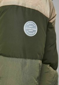 Kurtka puchowa w zielono-beżowym wzorze. Posiada okrągłą naszywkę z logo z napisem, gładki materiał oraz kontrastowe bloki kolorów.