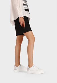 Sneakers bianchi con una superficie testurizzata e un accento argento sul tallone, abbinati a una gonna nera e una camicia bianca con motivi geometrici.