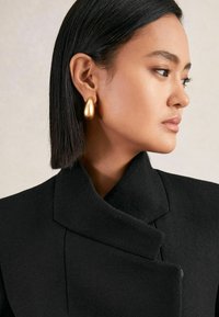 Cappotto di lana nera con colletto alto, abbinato a un grande orecchino a goccia in oro. Texture liscia e silhouette elegante, che mette in mostra un design contemporaneo.