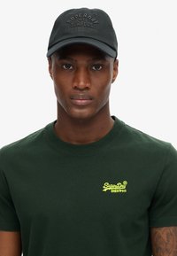 T-shirt in cotone verde scuro con maniche corte, caratterizzato da un piccolo logo giallo ricamato. Cappellino nero con logo in rilievo e visiera curva.