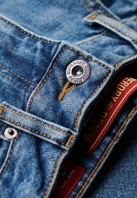 Jeans de mezclilla en un color azul claro, con costuras amarillas en contraste, un botón metálico con la marca "Superdry" y etiquetas internas visibles.