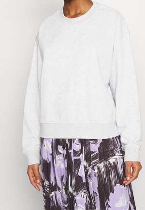 Personne portant un sweat-shirt gris clair à manches longues et une jupe plissée avec des motifs abstraits de coups de pinceau noirs et violets.