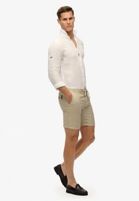 Camisa de lino ligera de botones en blanco, combinada con pantalones cortos beige con cintura ajustable y bolsillos laterales, llevada con mocasines oscuros.