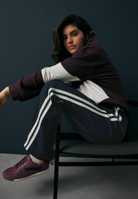 Pull à manches larges maroon et blanc avec un pantalon de survêtement marine, à rayures blanches. Baskets bordeaux. Modèle assis sur une chaise noire.