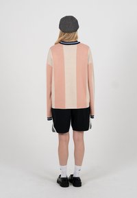 Osoba w szarej czapce, długim swetrze z szerokimi pionowymi różowymi i beżowymi paskami, czarnych krótkich spodenkach, białych skarpetkach i czarnych butach, stojąca tyłem.