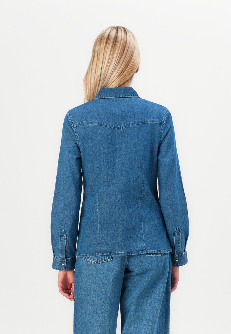 Chaqueta de mezclilla en azul medio, con cuello de solapa, mangas largas con puños abotonados y una silueta ajustada. Tejido texturizado con costuras traseras.