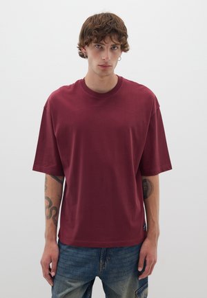 Camiseta oversize burdeos de algodón con mangas cortas, cuello redondo y una pequeña etiqueta de la marca en el lateral; combinada con jeans azules.
