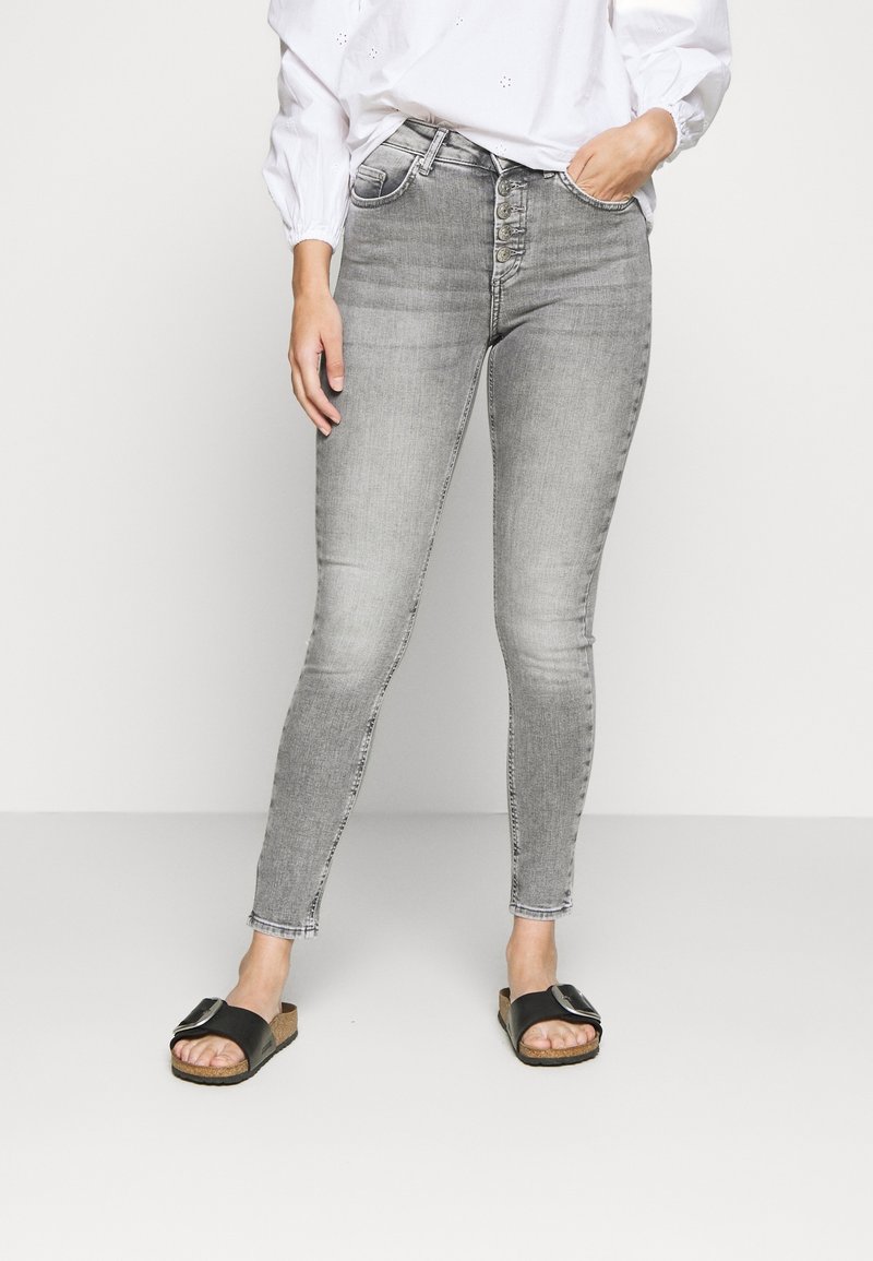 Jeans de mezclilla gris ajustados con cintura alta, cierre de cinco botones y un ligero acabado desgastado. Combinados con sandalias negras con una correa con hebilla.