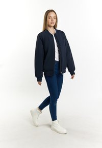 Marine bomberjack met geribbelde manchetten en kraag, gecombineerd met een wit topje en een blauwe strakke jeans. Model draagt witte hoge sneakers.