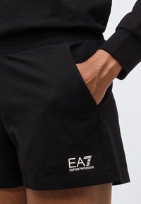 Persona con pantalones cortos negros EA7 Emporio Armani con la mano en el bolsillo sobre un fondo claro y liso.