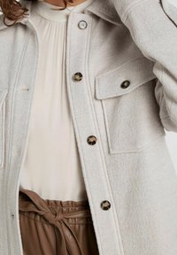 Jasnoszara kurtka o fasonie shacket, wykonana z mieszanki wełny, z dwoma kieszeniami na piersiach, czarnymi guzikami i luźnym krojem, nałożona na bluzkę w neutralnych odcieniach i brązowe spodnie.