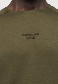 Άτομο με σκούρο δέρμα που φοράει πουκάμισο ελιάς με ανεβασμένο λογότυπο "Porsche BOSS" στο κέντρο του άνω στήθους.