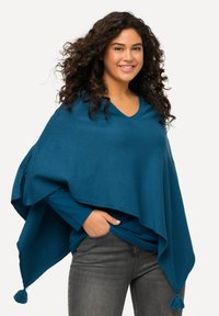 Poncho turchese con scollo a V, in tessuto lavorato a maglia, e frange decorative sull'orlo. Abbinato a una maglietta blu a maniche lunghe e jeans grigi.