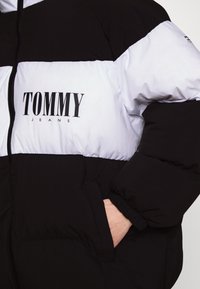 Czarna i biała kurtka puchowa zapinana na zamek, z dużym logo „TOMMY JEANS”, elastycznymi mankietami i boczną kieszenią.