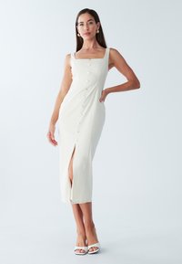 Robe midi couleur crème avec un col carré, des détails à boutons sur les côtés et une fente à l'avant. Caractérisée par des bretelles fines et une texture lisse.