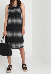Schwarz-weiß gestreiertes ärmelloses Kleid mit lockerer Passform, kombiniert mit einer schwarzen Tragetasche und schwarzen Espadrilles-Sandalen. Glatte Textur.
