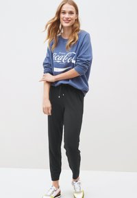 Modrá mikina s potiskem "Enjoy Coca-Cola", v kombinaci s černými tepláky s šňůrkou v pase; barevné sportovní boty dotvářejí outfit.