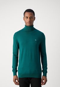 Pull à col roulé vert en tricot léger, avec un col et des poignets côtelés, et un petit logo sur la poitrine.