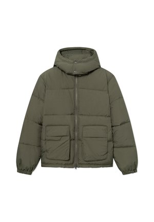 Homme debout les mains dans les poches, portant une doudoune verte, une casquette noire, un jean noir délavé et des chaussures noires sur fond blanc.
