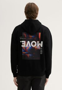 Felpa nera con cappuccio e stampa grafica sulla schiena che presenta forme astratte colorate e testo. Il tessuto sembra morbido e texturizzato.