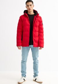 Chaqueta acolchada roja con capucha, con cierre de cremallera y puños elásticos. Combinada con una camiseta negra, vaqueros azul claro y zapatillas blancas.