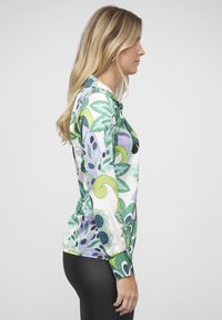 Halenka s dlouhým rukávem z lesklé látky, zdobená velkými květinovými vzory v zelené, fialové a bílé barvě, s úzkými černými rukávy a kulatým výstřihem.