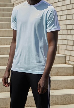 Homme portant un t-shirt Adidas bleu clair et un pantalon de survêtement Adidas noir, debout à l'extérieur près d'escaliers en béton et d'un mur en briques.