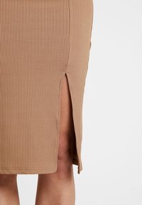 Beżowa spódnica prążkowana z bocznym rozcięciem odsłaniającym część nogi na tle białym.