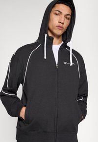 Sweat à capuche zippé noir avec capuche doublée, accents blancs et poches avant. Comprend un logo Champion sur la poitrine et des poignets côtelés.