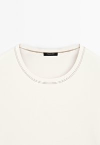 T-shirt a maniche corte di colore crema con scollatura rotonda, cuciture a contrasto e un'etichetta nera sul collo che riporta la dicitura "Massimo Dutti, made in Portugal."