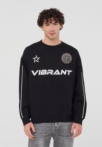 Felpa nera con collo rotondo, caratterizzata dalla scritta "VIBRANT" in grassetto bianco, un logo circolare e strisce bianche sui lati delle maniche.