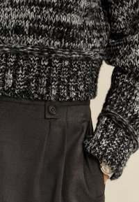 Chunky stickad tröja i svart och grått med ribbad nederkant, kombinerad med mörka byxor med knappdetalj och sidofickor. Silverarmbandet är synligt.