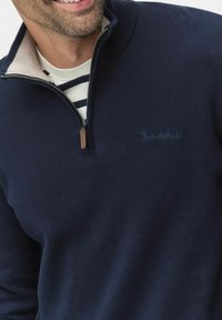 Marinblå polotröja med dragkedja tillverkad av mjukt material med en beige inre krage. Har en subtil logotyp och en texturerad yta.