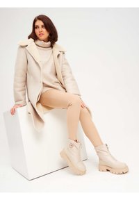 Abrigo acolchado beige con un cuello suave, suéter de punto crema con cuello alto, leggings color beige y botas claras con suela gruesa y cremallera lateral.