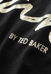 Melnā auduma materiāls ar zelta metālisku grafisko dizainu un tekstu "BY TED BAKER" treknrakstā, uzsverot gludu tekstūru.
